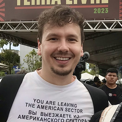 Foto do pesquisador André Lopes. Ele é um homem branco, com cabelo ondulado, castanho claro, barba e bigode. Na imagem, ele está sorrindo em frente a fachada do Acampamento Terra Livre